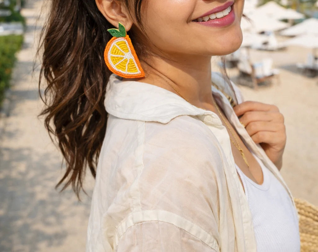 Orange beaded dangling earrings with green leaf accents, worn close-up