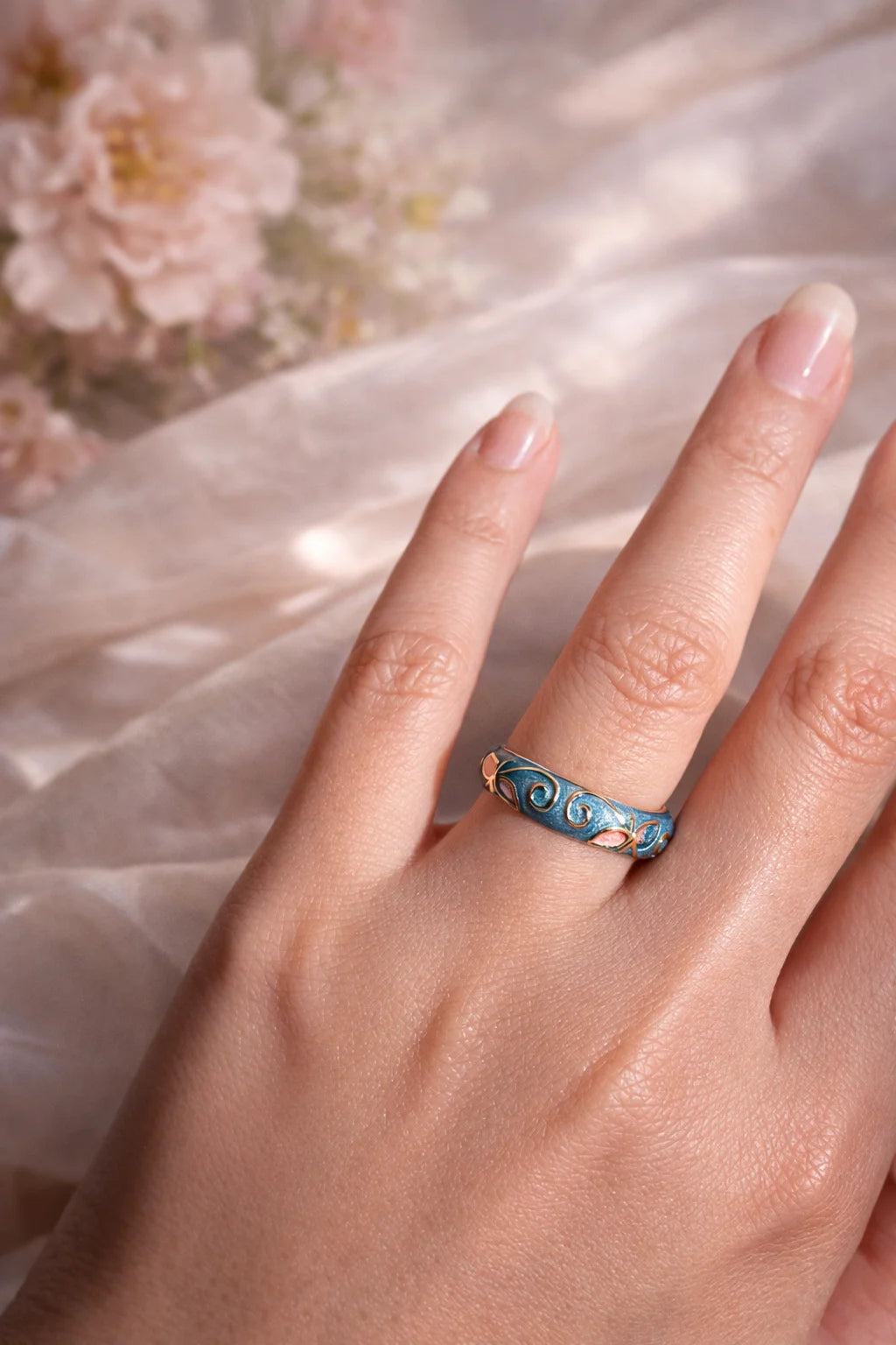 Blue enamel ring on finger with gold edge, scrollwork and pink insets on a slim band