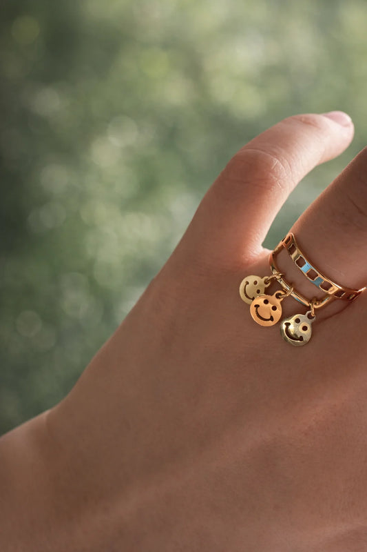 Gold-plated smiley charm ring with blue enamel square insets, worn on finger